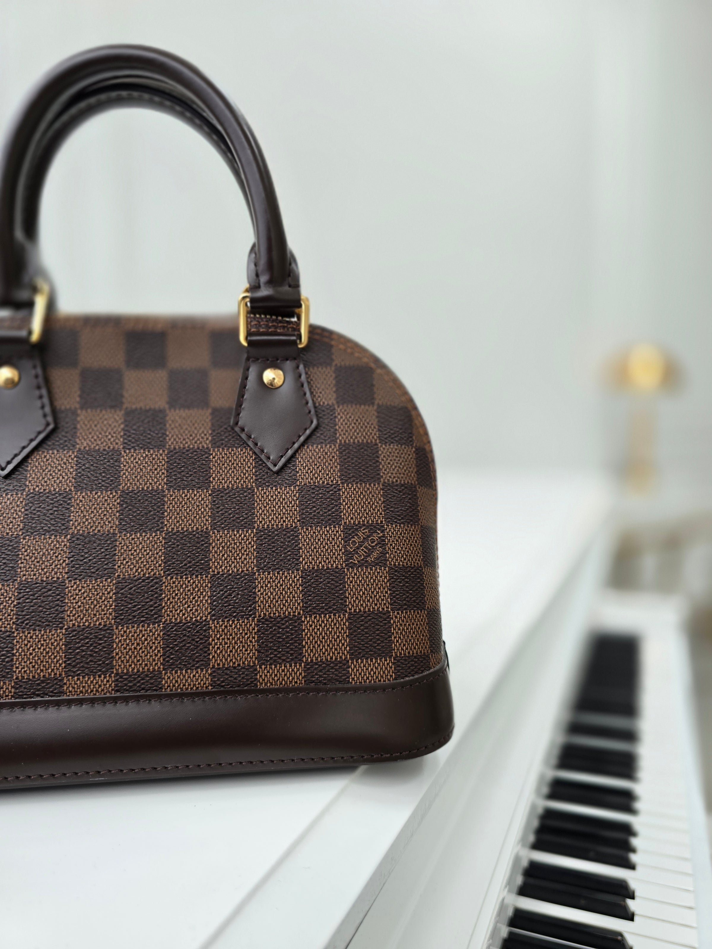 silhouette photo of a pre-owned louis vuitton alma bb in signature damier ebene brown canvas with dual rolled top handles on top of a white grand piano